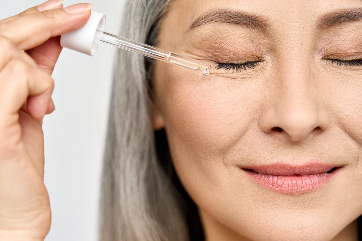 Ältere Frau mit grauen langen Haaren und geschlossenen Augen tropft mit einer Pipette Serum auf ihre Wange